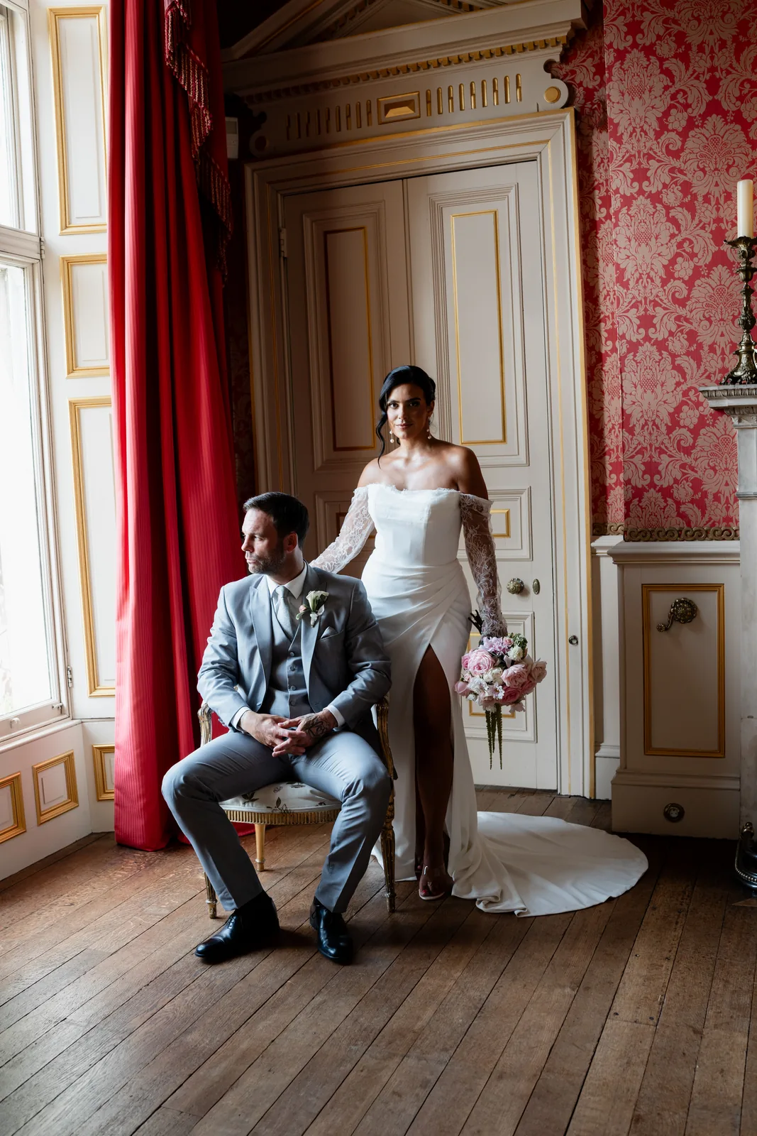 Groom seated and bride standing beside him in an ornate red drawing room